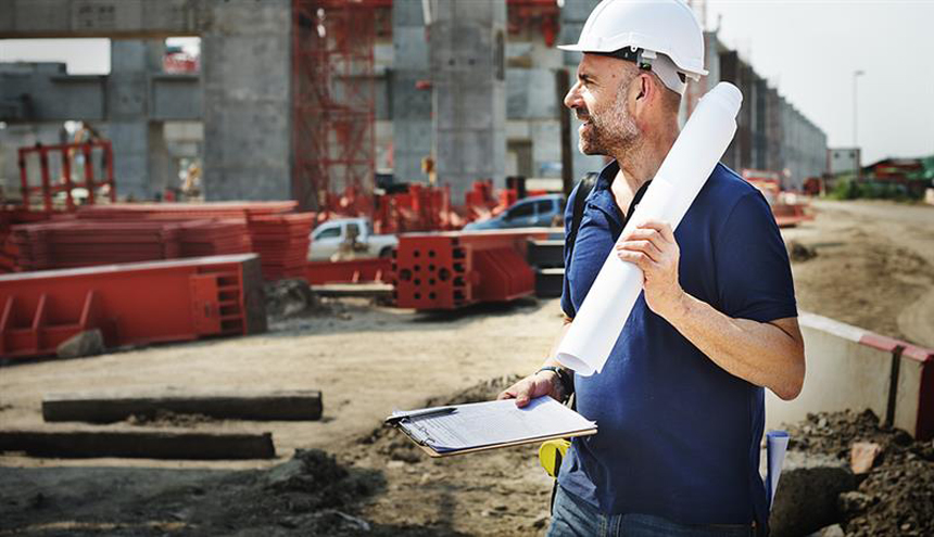 A construction worker wearing a white hard hat and blue shirt holds rolled blueprints and a clipboard, standing at a building site.