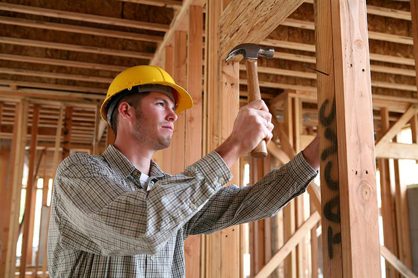 Construction worker wearing a yellow hard hat and checkered shirt, using a hammer to drive a nail into a wooden frame at a building site.