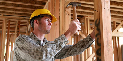 Construction worker wearing a yellow hard hat and checkered shirt, using a hammer to drive a nail into a wooden frame at a building site.